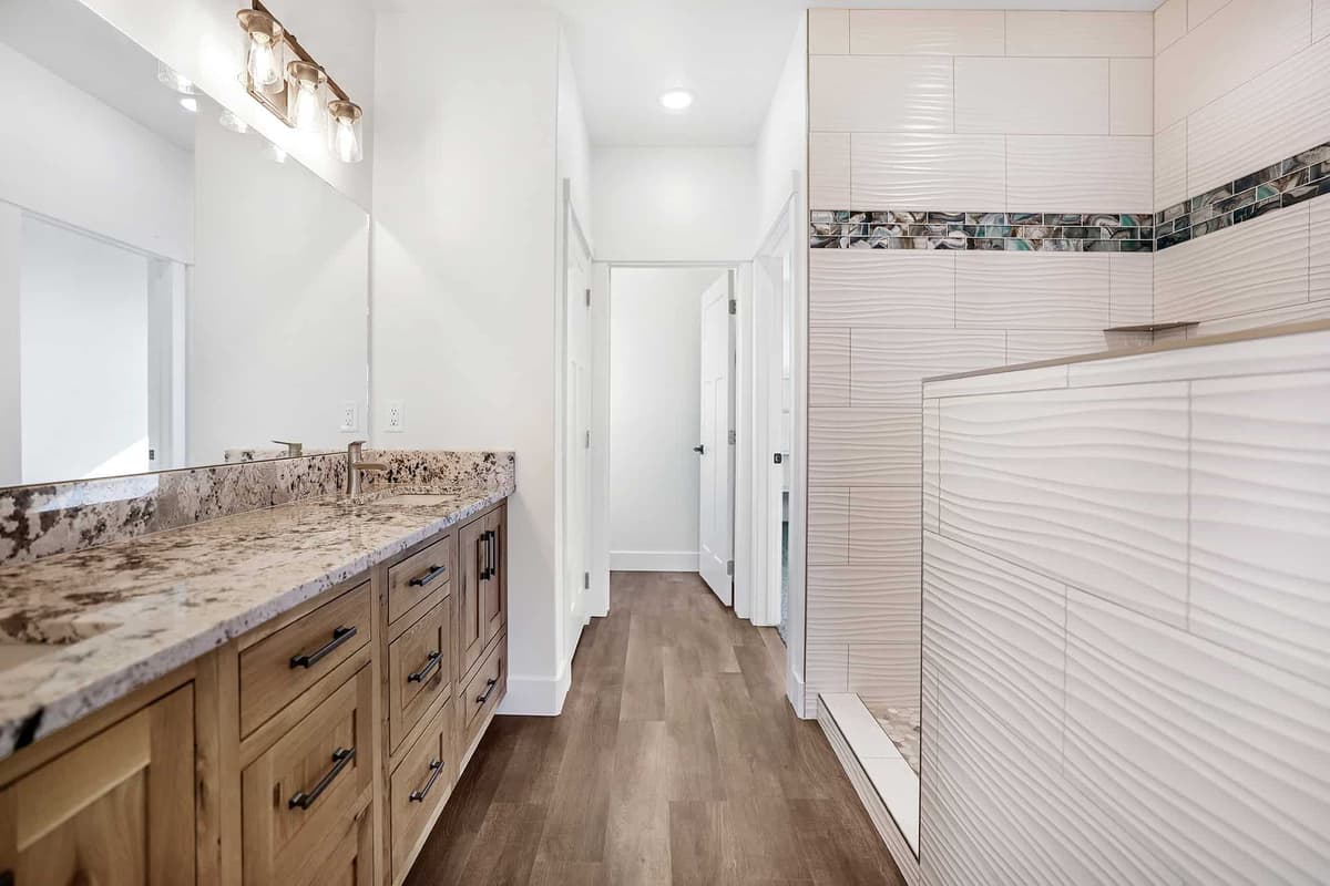 A bathroom with a granite-topped wood vanity and a walk-in shower tiled in large wavy white tile with a glass accent band